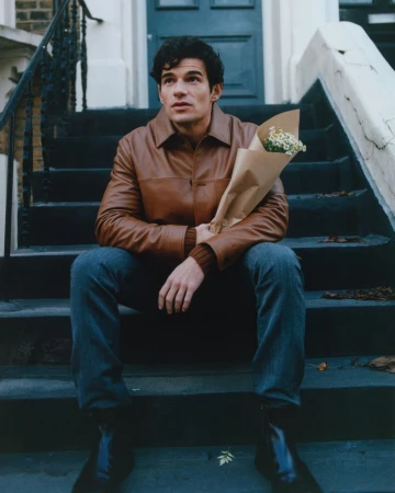 A young adult male with dark curly hair sits on stone steps holding a small bouquet of white flowers, wearing jeans and a brown leather jacket.