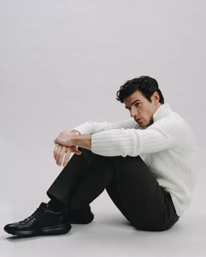 Man wearing a white ribbed turtleneck and dark trousers sitting against a plain white studio background