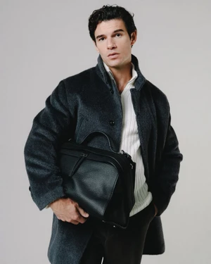 Actor Paul Forman wearing a dark wool overcoat and white knit sweater while holding a black textured leather bag.