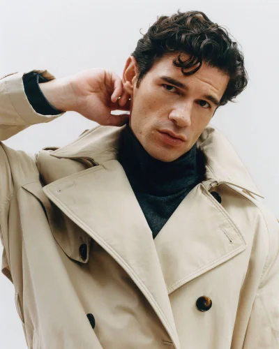 Paul Forman wearing a beige trench coat and dark turtleneck sweater against a plain white studio background.