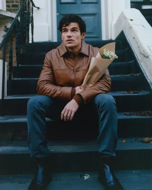 Paul Forman sitting on outdoor stone steps wearing a brown leather jacket and holding a bouquet of flowers in kraft paper.