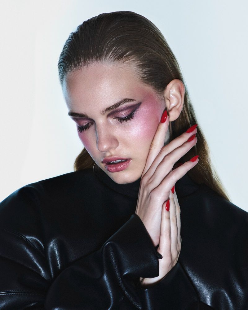 Close-up portrait of a woman with dark pink eye makeup, red nail polish, and slicked-back hair posing in a black leather jacket in a studio.