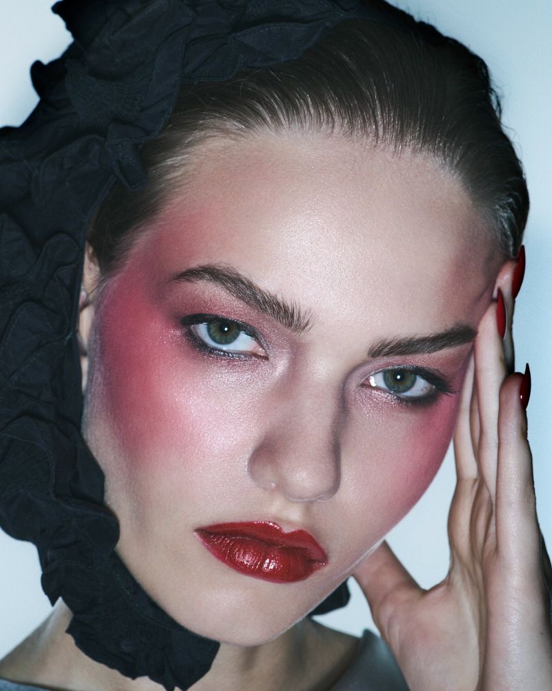 Close-up portrait of a woman with dramatic makeup, styled with dark fabric, and resting her fingers against her temple in studio lighting.