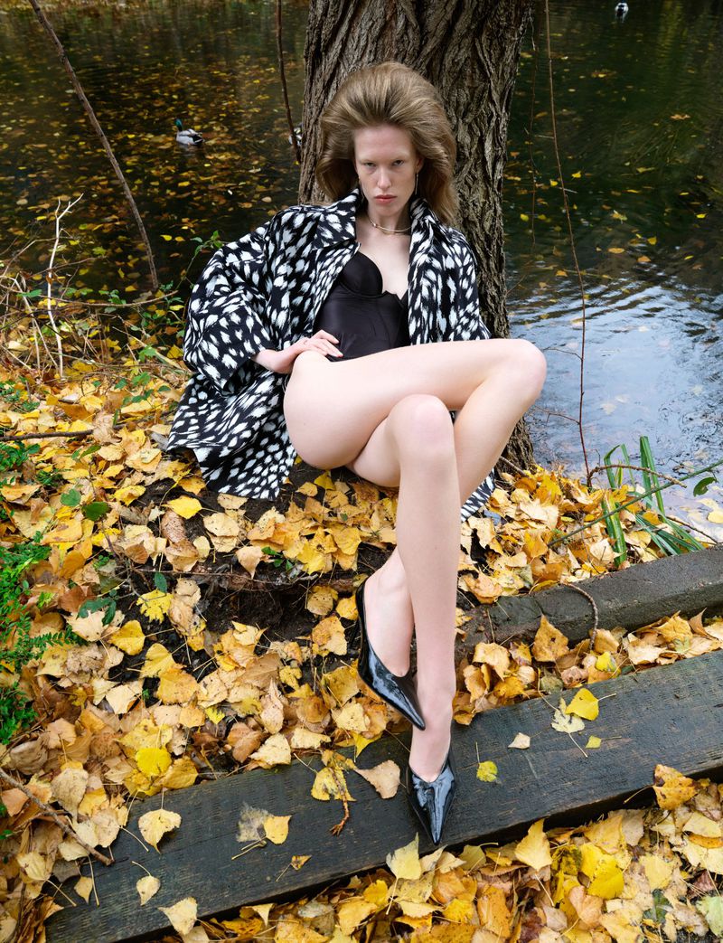 A woman in a black and white jacket poses by a tree near water. Autumn leaves cover the ground. She wears black heels.