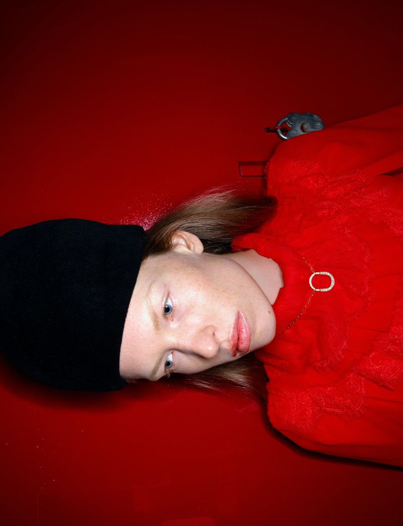 Portrait of an adult female lying on a red surface, wearing a black beanie and red sweater.  She is also wearing a necklace.