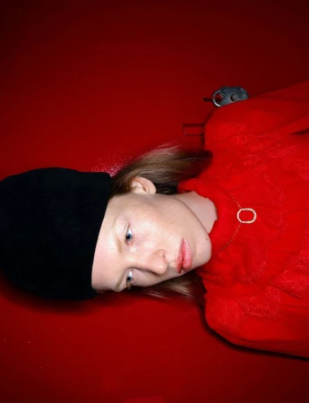 Portrait of an adult female lying on a red surface, wearing a black beanie and red sweater.  She is also wearing a necklace.