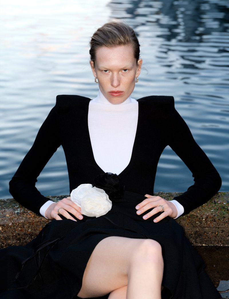 Woman in black dress and white turtleneck poses outdoors on concrete ledge with waterfront background, demonstrating fashion and style.