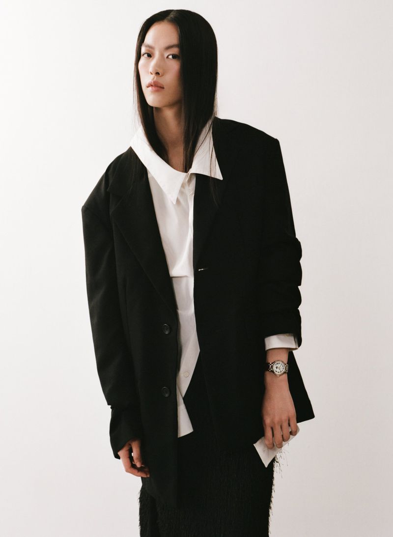 East Asian woman posing in an oversized black blazer and white collared shirt against a white seamless studio backdrop.