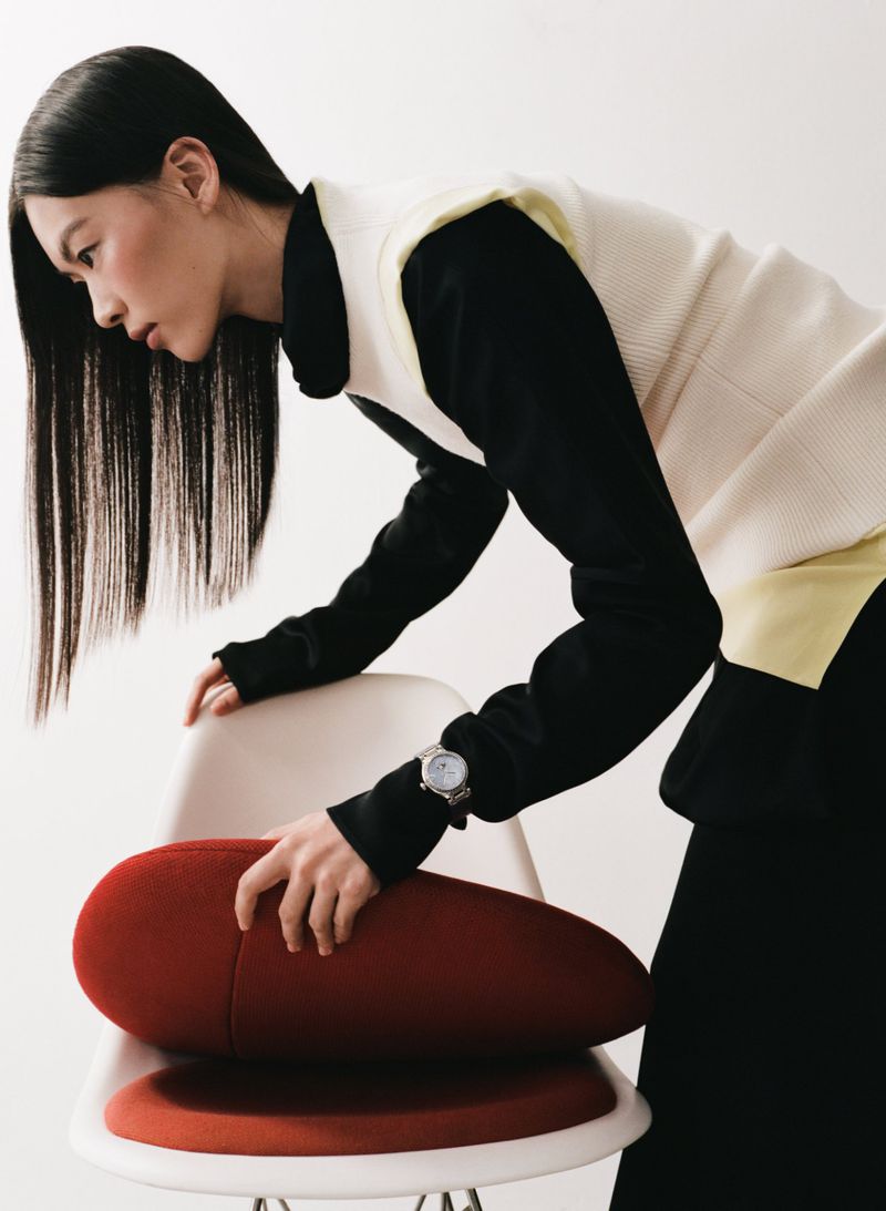 Woman in sleeveless sweater leans over chair with red cushion in minimalist indoor setting, styled in neutral colors with long black hair.