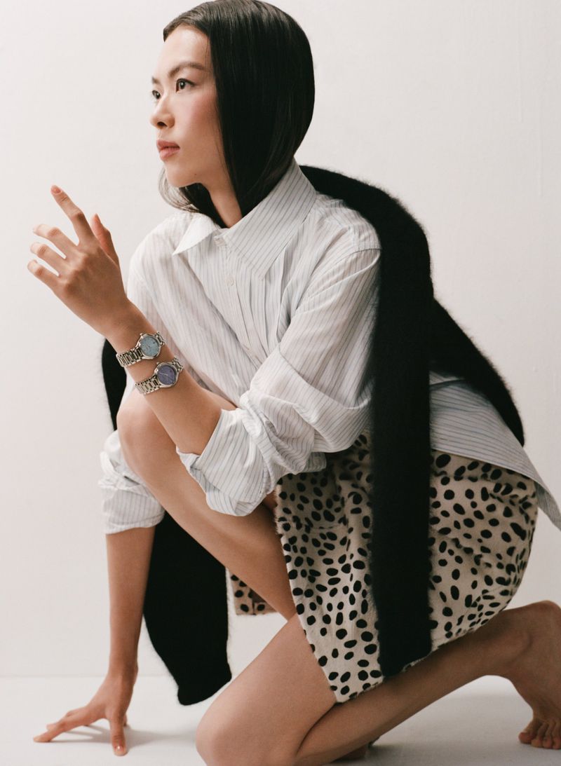 An Asian woman in a light-striped shirt poses in a crouched position wearing two bracelets and a dalmatian-print skirt against a white background.