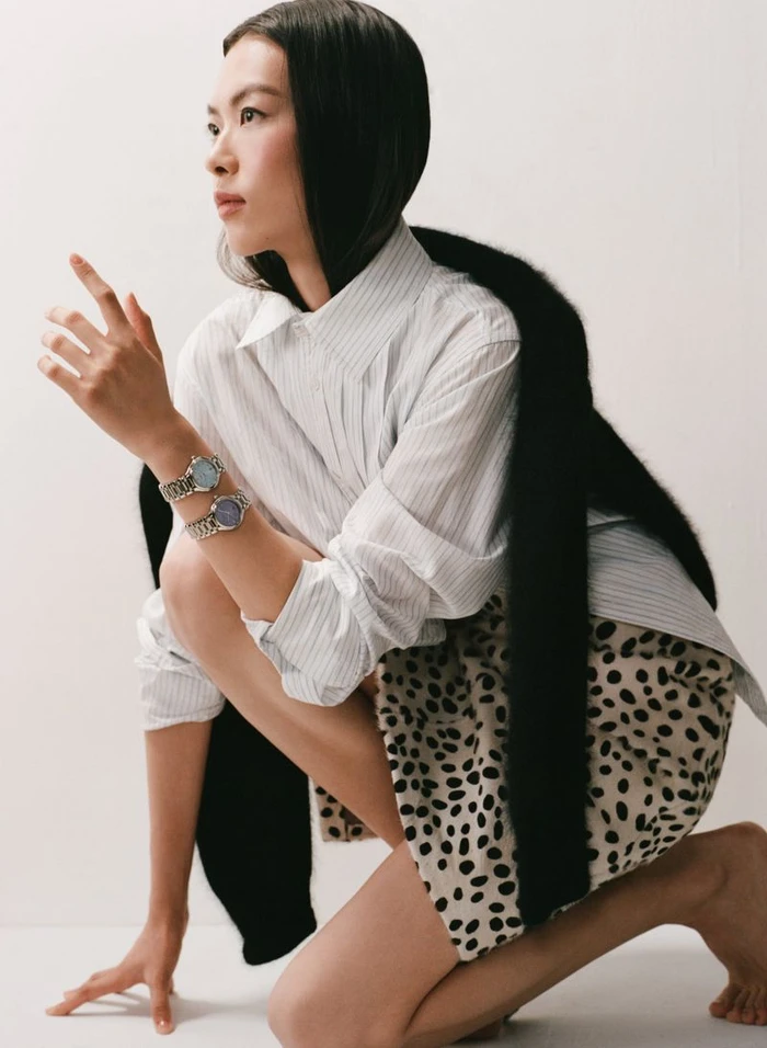 East Asian woman crouching in a white striped shirt, spotted skirt, black faux fur stole, and two wristwatches against a white backdrop.