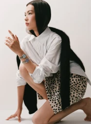 East Asian woman crouching in a white striped shirt, spotted skirt, black faux fur stole, and two wristwatches against a white backdrop.