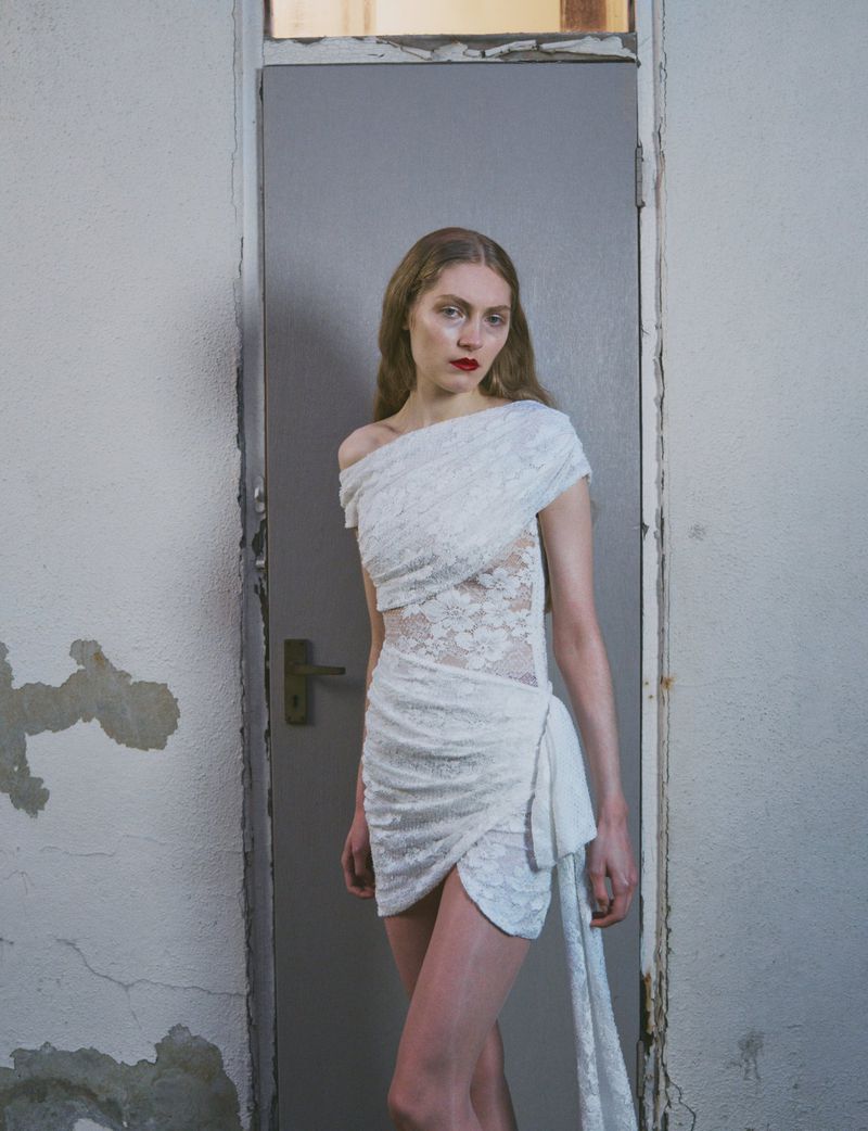 A poised woman in a short white lace dress stands indoors by a grey door, highlighted with red lipstick and soft lighting for fashion.
