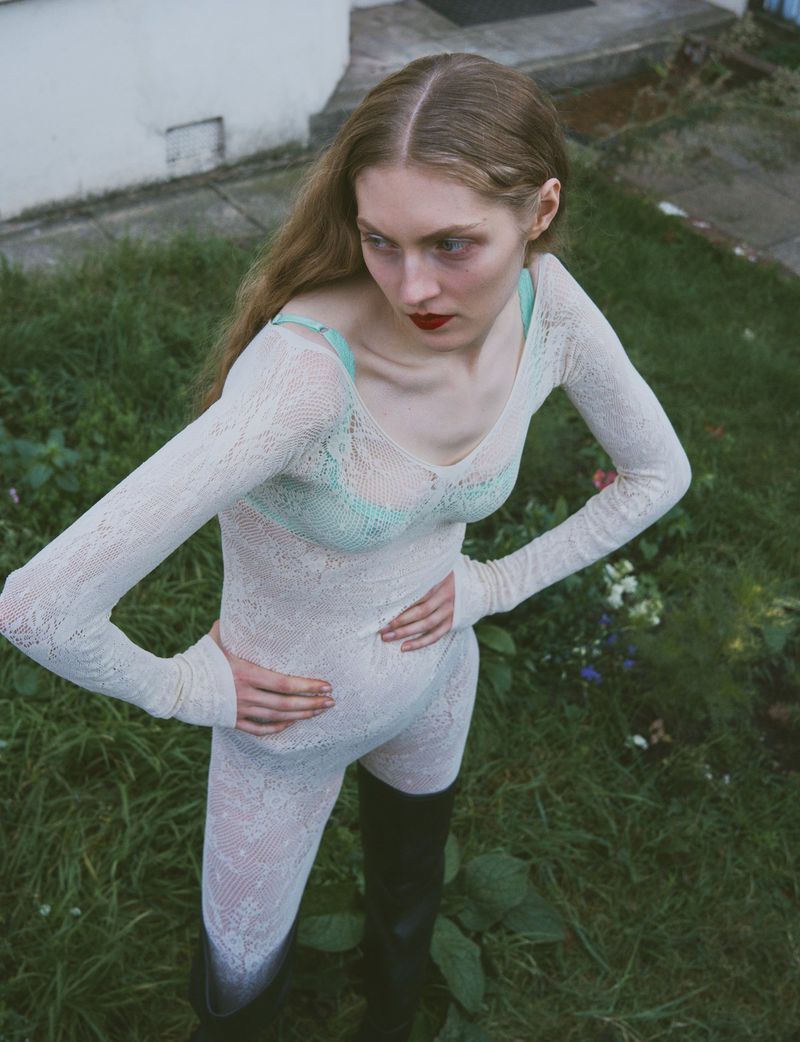 Blonde woman in a textured bodysuit posing in a grassy outdoor setting near a house, wearing dark boots and red lipstick.