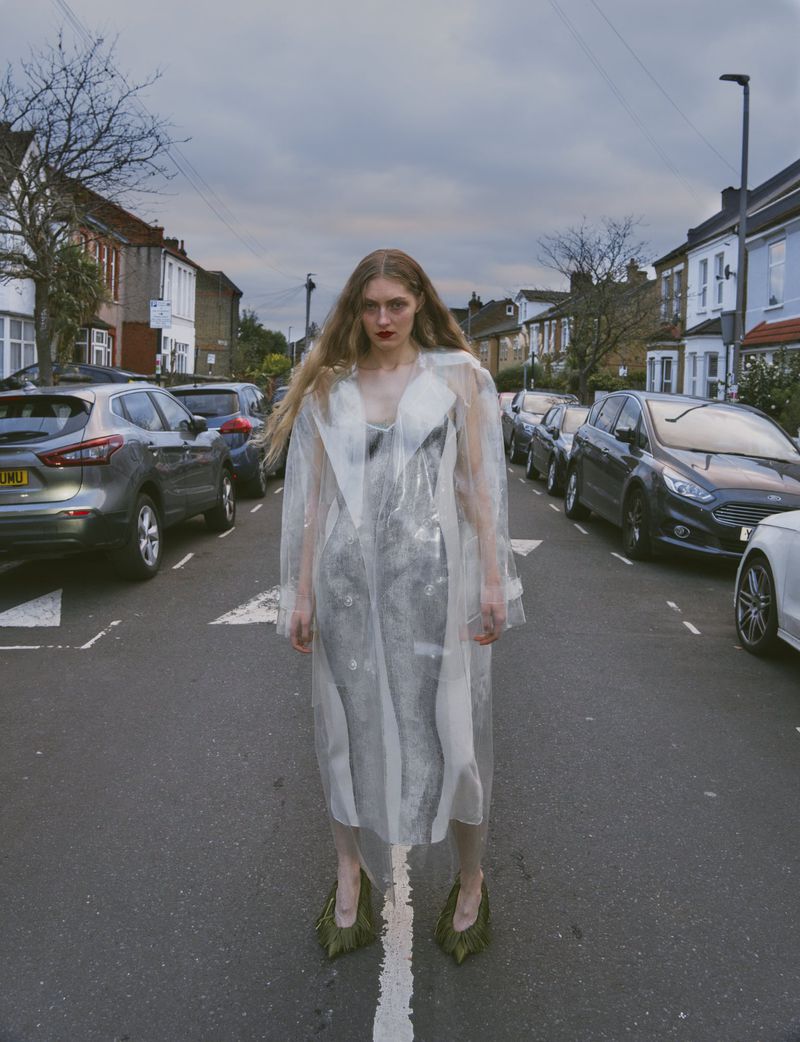 Fashion portrait of a long-haired woman in a transparent coat and green heels, standing on a street with parked cars during the daytime.