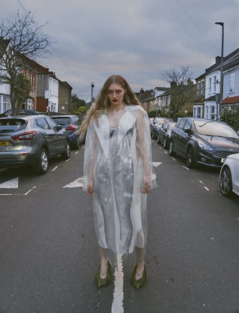 Fashion portrait of a long-haired woman in a transparent coat and green heels, standing on a street with parked cars during the daytime.