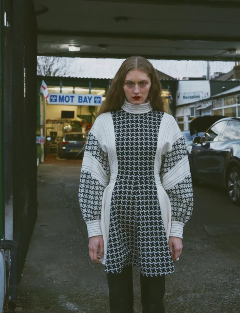 A long-haired woman in a patterned dress stands in front of an auto shop bay; cars are in the background. The lighting is diffused.