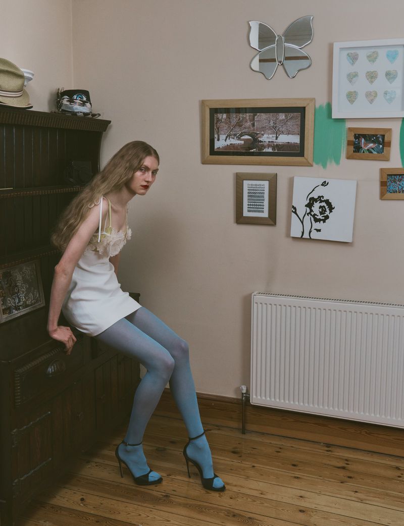 A slender woman in a white dress, blue tights, and black heels poses indoors next to a dark wood cabinet with decorative wall art.