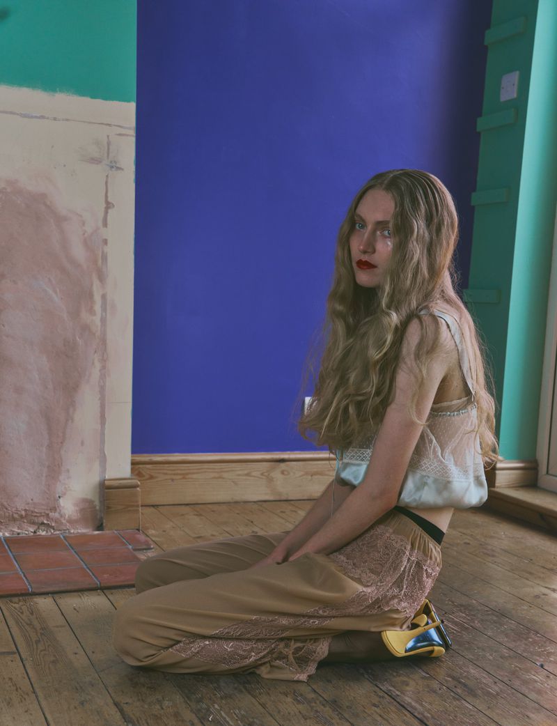 Model Constanze Van Rosmalen kneeling on wooden floorboards wearing a sheer lace top and beige trousers in front of a blue wall.