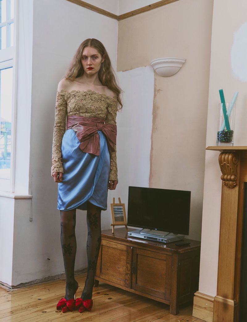 A slender woman with long brown hair poses indoors wearing a tan lace top, blue satin skirt, red shoes, and dark tights.