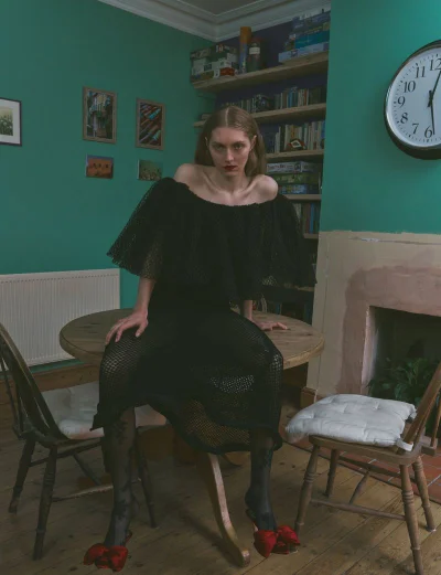 Model in an off-the-shoulder black dress and red bow shoes sitting on a round wooden table in a green room with a fireplace.
