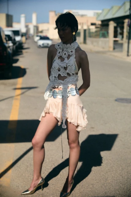 A fashionable young woman with short dark hair poses in an artful outfit on an outdoor street in the daylight, casting a strong shadow.