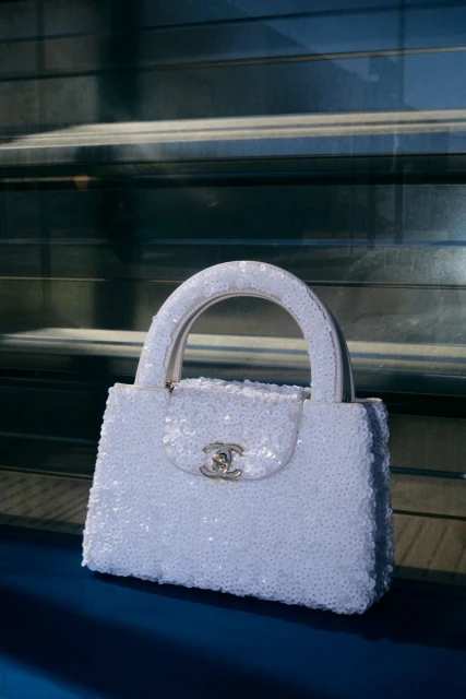 Elegant white Chanel handbag with a top handle and silver logo, placed on a reflective blue surface with a blurred gray background.