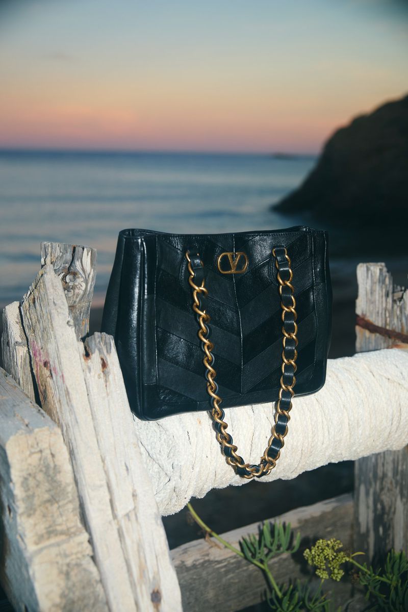 Valentino Garavani Locò small chain shoulder bag with VLogo Signature, black chevron leather, gold-tone chain, and sunset beach backdrop.