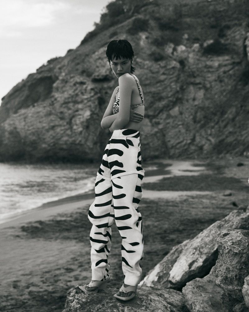 Monochrome shot of a person in a bikini top and patterned pants posing on a rock formation with a natural rocky beach background.