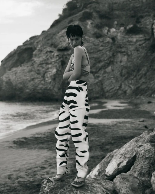 Monochrome shot of a person in a bikini top and patterned pants posing on a rock formation with a natural rocky beach background.