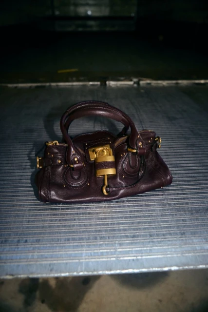 Dark purple leather satchel handbag with gold-toned padlock and hardware on a ribbed metal surface, featuring a structured design.