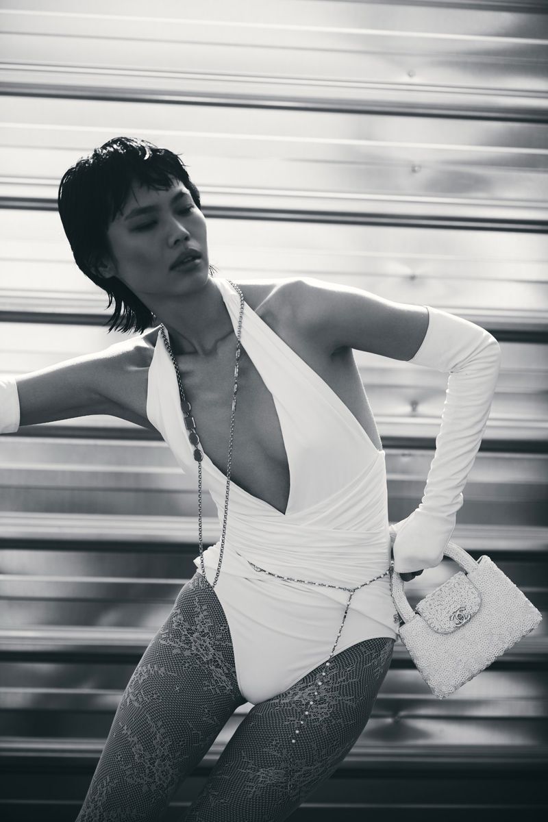 Fashion shot of an adult woman wearing a swimsuit, gloves, hosiery, and jewelry, holding a handbag, posing indoors.