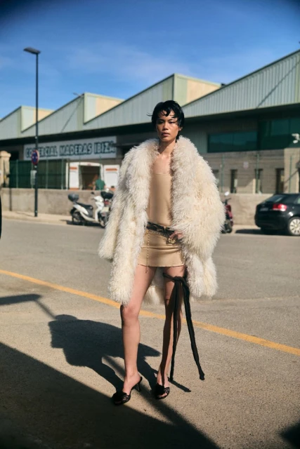 Fashionable person in a fluffy coat and mini dress poses on a city street with industrial buildings and a scooter visible in the background.