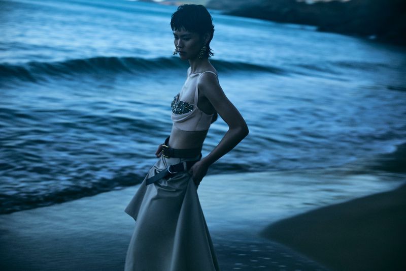 A slender woman stands on a sandy beach in a fashionable light pink top and khaki skirt. Waves roll behind her under a twilight sky.