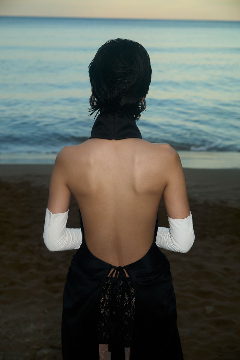 Back view of woman in open-back dress on beach at sunset. The dress has lace detailing, and the sky is a soft blend of colors.