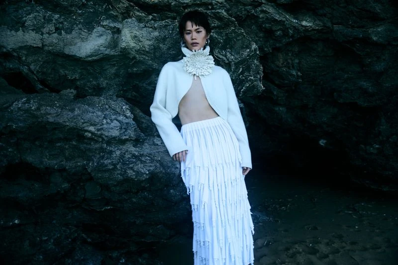Woman poses in white skirt and cropped jacket with large floral collar in front of a dark rock formation on a beach.