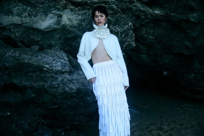 Woman poses in white skirt and cropped jacket with large floral collar in front of a dark rock formation on a beach.