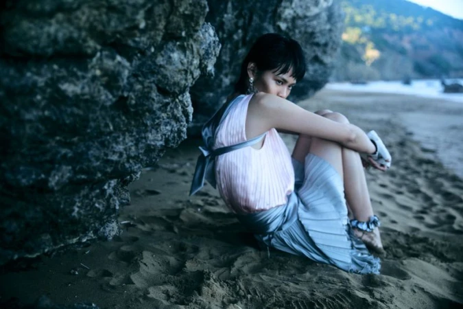Model Xia Yuancen seated in the sand beside a dark rock formation wearing a pleated pink top and pale blue skirt.