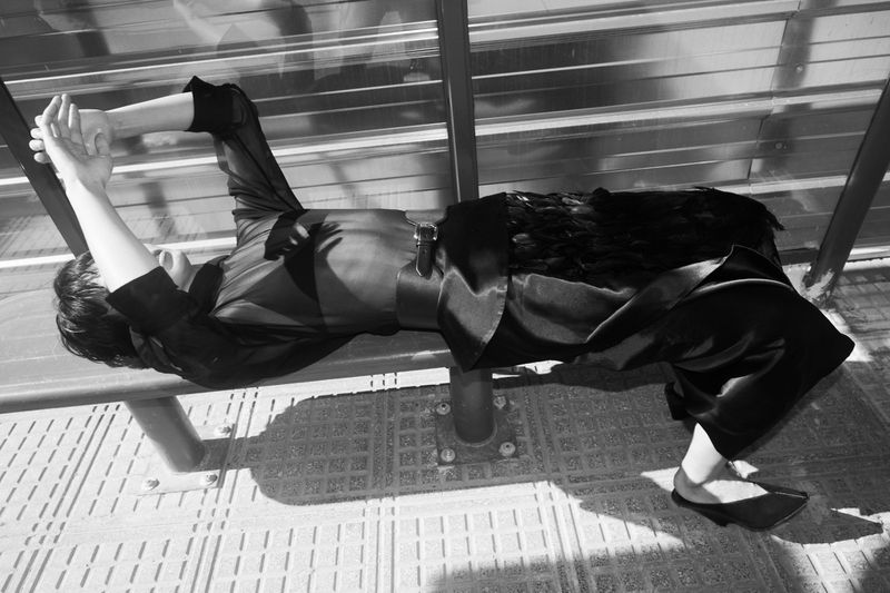 Monochrome fashion shot of a woman lying on a bench in a black outfit with feathered detailing, set against a backdrop of sunlight and shadows.