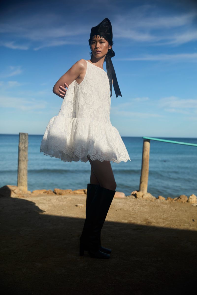 A fashion shot of a young adult woman in a white dress, black boots, and a black headpiece, posing against a seaside background.