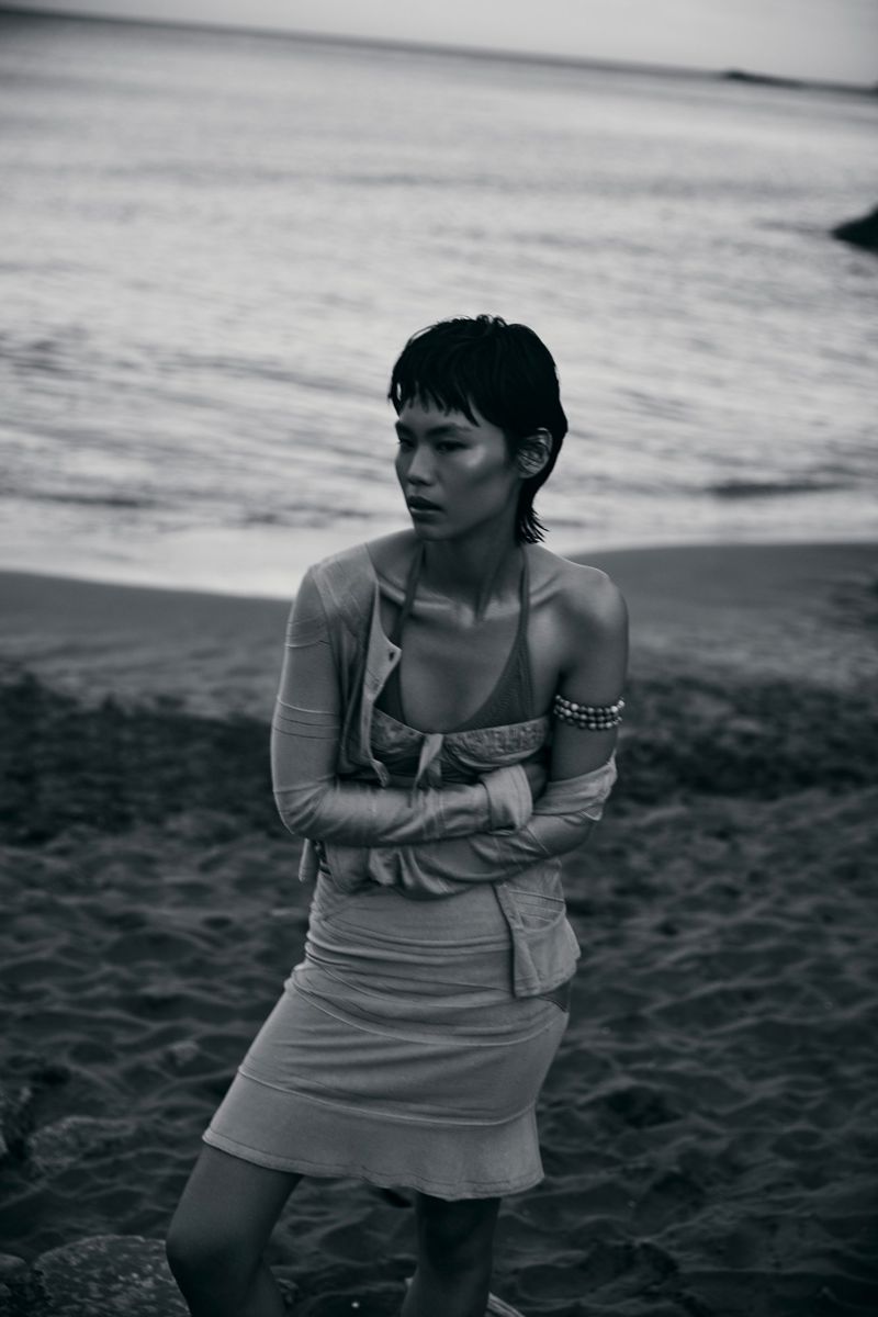 Model Xia Yuancen with short hair wearing a layered top and skirt on a sandy beach shoreline in monochrome.