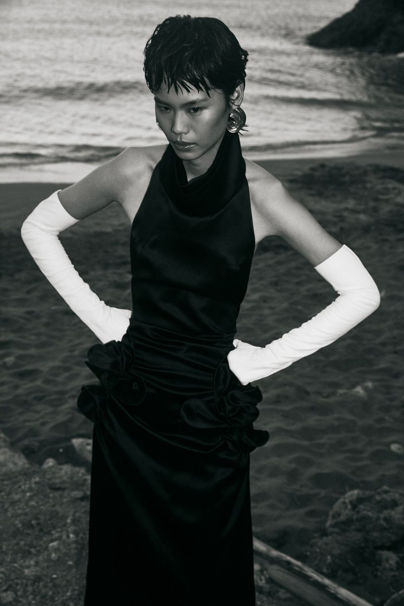 Model Xia Yuancen posing in a black halter dress with long white opera gloves on a rocky coastal shoreline.