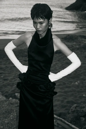 Model Xia Yuancen posing in a black halter dress with long white opera gloves on a rocky coastal shoreline.