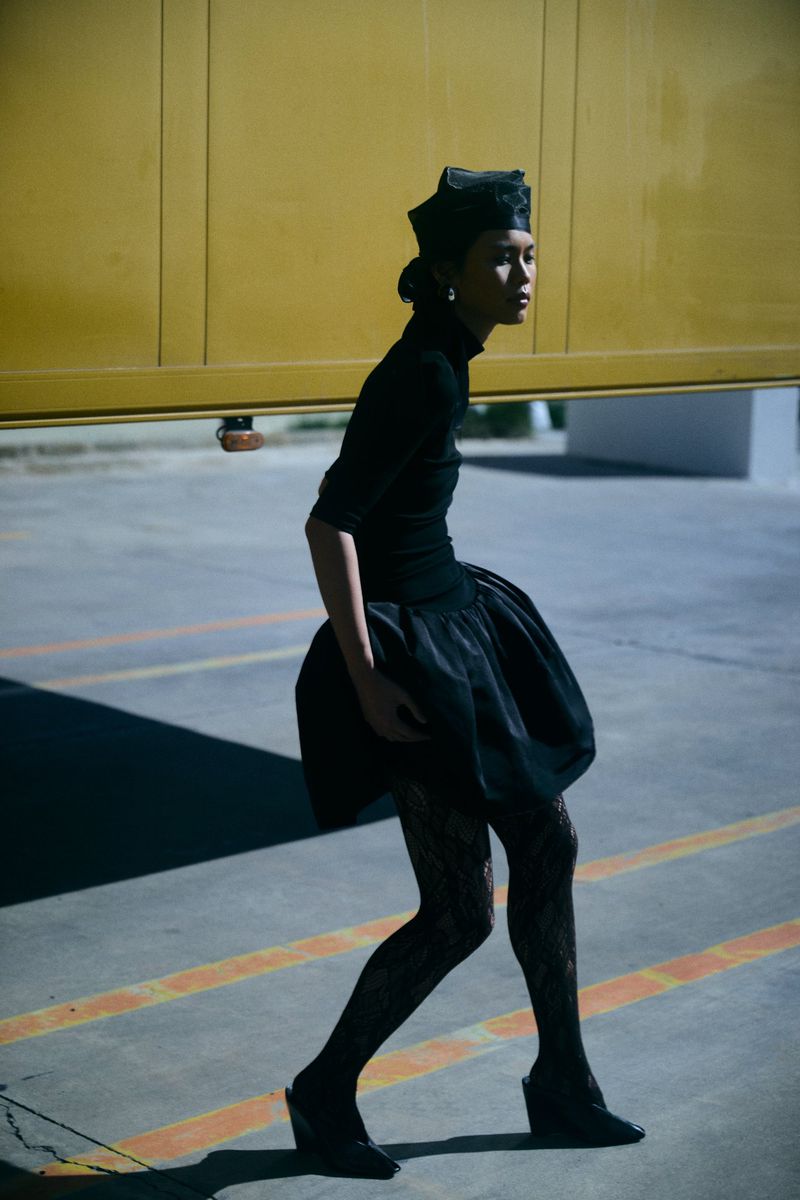 Elegant female model in black formalwear and patterned tights walks across an empty parking lot with a yellow garage door behind her.