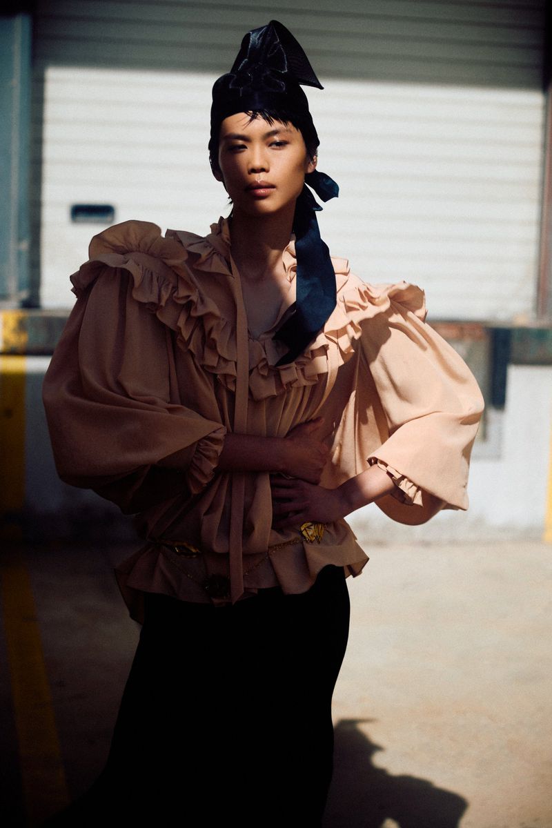 Fashion editorial shot of a person posing outdoors in a beige ruffled blouse, black skirt, and black headpiece in an urban setting.