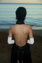 Model from behind wearing a black backless dress and white gloves standing facing the ocean on a sandy beach at dusk.