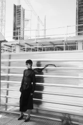 Model Xia Yuancen in a black belted dress and sunglasses standing before a corrugated metal wall with a scarf floating in the wind.