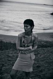 Model Xia Yuancen with short hair wearing a layered top and skirt on a sandy beach shoreline in monochrome.