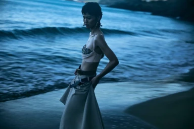 Model Xia Yuancen poses at the water's edge on a beach in an embellished cropped top and draped skirt at dusk.