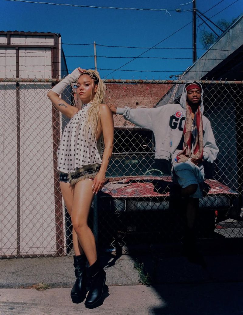 Two fashionably dressed people, a woman and a man, pose in an urban setting with a chain-link fence and a rusty car in the background.